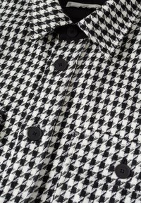 Tissu à motif pied-de-poule noir et blanc avec des boutons noirs et un col, comprenant une poche poitrine et une texture tissée.