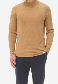 Brown turtleneck sweater in soft knit fabric, featuring long sleeves and a fitted design. Worn over dark pants with a small brand tag visible.