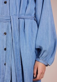 Ljusblå denimklänning med insvängd midja, knäppning framtill och lösa, rynkade ärmar. Har ett matchande bälte i samma tyg.