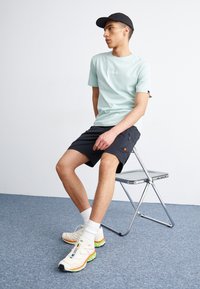 Lys grønn bomull T-skjorte med logo, kombinert med svarte shorts og multicolor joggesko. Modellen sitter på en krom sammenleggbar stol.