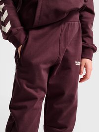 Barn iført mørkelilla træningsbukser med matchende sweatshirt, den ene hånd i lommen, hvide Chevron-striber på ærmet og logo på bukserne.