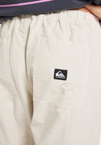 Beige cargo shorts yang terbuat dari bahan ringan, dilengkapi dengan pinggang elastis, saku samping, dan patch logo Quiksilver hitam di belakang.