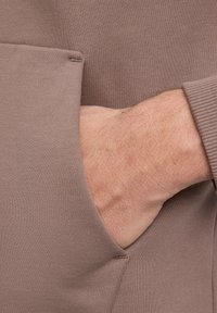 Hoodie en tissu marron avec une poche kangourou. Matériau texturé et doux avec des accents côtelés près de la poche. Main positionnée à l'intérieur de la poche.
