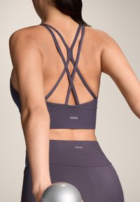 Ljupka sportska grudnjak ljubičaste boje s trakama koje se križaju na leđima, izrađen od glatkog materijala. Naziv marke "OYSHO" vidljiv je na traci i pojasevima.