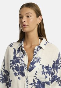 Ritratto di una donna con lunghi capelli castani, che indossa una camicia a fiori con motivi blu su uno sfondo chiaro. Guarda di lato.