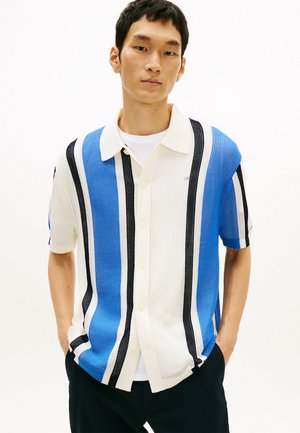 Ung mand iført en kortærmet skjorte med lodrette blå, hvide og sorte striber, stående med hænderne i lommerne.