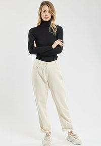 Frau, die mit verschränkten Armen vor einem weißen Hintergrund steht, trägt einen schwarzen Rollkragenpullover, beigefarbene High-Waist-Hosen und weiße Sneakers.