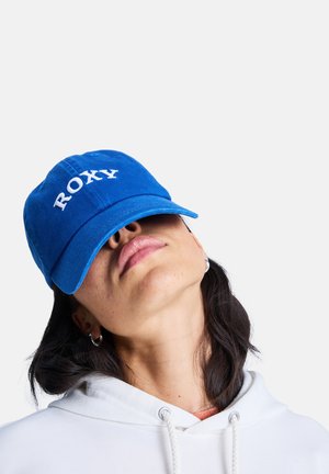 Persoon met een blauwe pet met de tekst "ROXY", een witte hoodie en zilveren oorringen, die het hoofd naar achteren kantelt tegen een witte achtergrond.