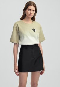 T-shirt ombre olive avec des manches courtes, présentant un patch en forme de cœur texturé, associé à une mini-jupe noire ajustée. Matériaux lisses et modernes.