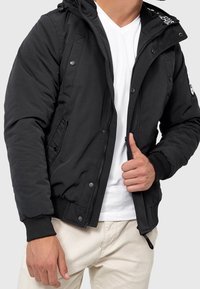 Zwarte zip-up jas met capuchon, met zijzakken, geribbelde manchetten en een soepele textuur. Draagt over een wit T-shirt.
