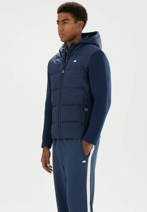 Joven con una chaqueta con capucha acolchada azul marino y pantalones deportivos azul marino a juego con rayas blancas en los laterales, de pie con las manos en los bolsillos.