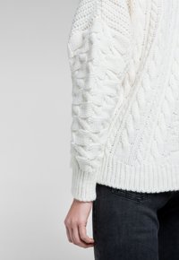 Vit kabelstickad tröja med ribbade muddar och fåll, som har strukturerade mönster längs ärmarna och en avslappnad passform.