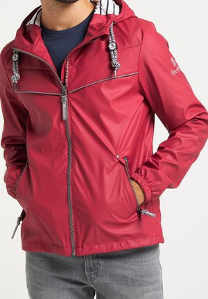 Mand iført rød hættejakke med hvidt stribet for og sorte og hvide snore, hænder i lynlåslommer på siden.