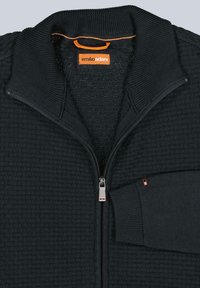 Schwarzer Sweater mit Reißverschluss, strukturiertem, gesteppte Muster, geripptem Kragen und Bündchen sowie einem orangefarbenen Akzent am Etikett und am Reißverschlusszug.