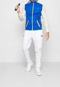 Blå zip-up jacka med vita ärmar och mönstrade detaljer, parat med vita träningsbyxor och vita sneakers, håller en tennisklubba.