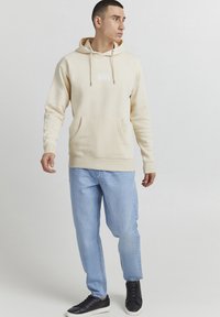 Felpa color crema con tasca frontale, chiusura con lacci e design testuale bianco. Indossata con jeans azzurri e sneakers nere.