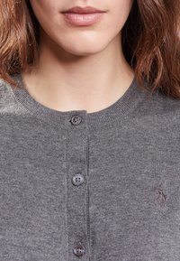 Harmaa neuletakki, joka on valmistettu pehmeästä neuloskankaasta, jossa on pyöreä pääntie, neljä nappia ja pieni kirjailtu logo vasemmassa rinnassa.