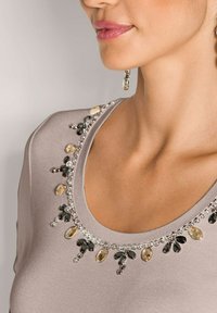 Vrouw in een beige top met een ronde halslijn, versierd met afwisselend zwarte en lichtbruine druppelvormige kralen en zilveren ronde kralen.