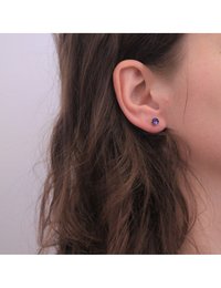 Boucles d'oreilles en argent avec des clous violets, présentant un design de gemme facettée et ronde. Les cheveux tombent légèrement sur l'oreille. Simple et élégant.