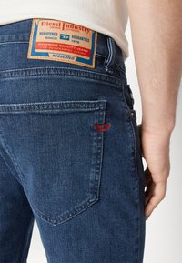 Jeans de mezclilla azul oscuro con un bolsillo trasero plano y un logo bordado en rojo. Presentan un parche de cuero marrón en la cinturilla con los detalles de la marca.
