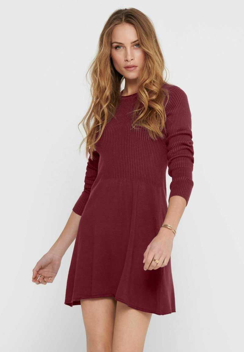 Robe en tricot bordeaux avec des manches longues côtelées et un corsage ajusté, s'élargissant légèrement à l'ourlet. Texture douce, style décontracté.