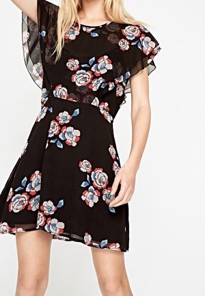 Žena v černých šatech s růžovými a modrými květinovými vzory a průhlednými volánovými rukávy na bílém pozadí.