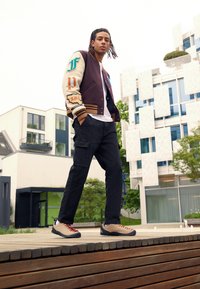 Maroon and cream varsity jacket with letter patches, black cargo pants, beige and red lace-up shoes, posed on wooden decking outside.