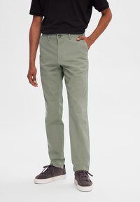 Chinos verde claro con un diseño de pierna recta, hechos de material de algodón, con cierre de botón y bolsillos laterales. Llevados con zapatillas de ante grises.