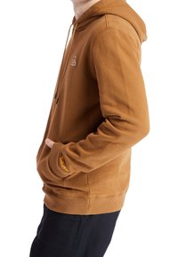 Persona vistiendo una sudadera marrón de Timberland con las manos en el bolsillo delantero, combinada con pantalones negros, mostrada de lado sobre un fondo blanco.