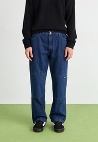 Denim trousers in dark blue with a relaxed fit, featuring seam detailing, pockets, and a small logo on the thigh. Worn with a black sweater.