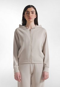 Sudadera beige con cremallera, capucha y mangas largas. Fabricada con tela suave, con bolsillos laterales y un ajuste relajado. Adecuada para uso casual.