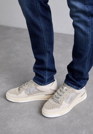 Beige tenisky s akcenty z perforované síťoviny a semiše, s perforovanými špičkami, plochými tkaničkami a bílou podrážkou. Kombinované s tmavě modrými džíny.