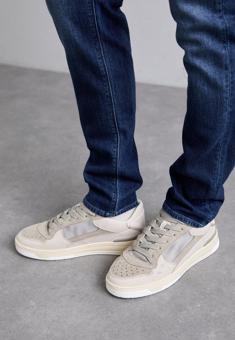 Sneaker beige con accenti in rete e suede, dotati di punte perforate, lacci piatti e suola bianca. Abbinati a jeans blu scuro.
