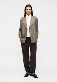 Femme portant un blazer croisé marron, une chemise blanche à col, un pantalon noir à jambes larges et des mocassins noirs, debout devant un fond uni.