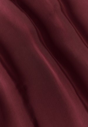 Mujer con un vestido largo de satén burdeos con escote en V pronunciado y detalle de lazo en el frente, combinado con tacones negros de tiras y pendientes dorados.