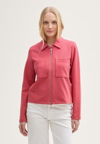 Veste rouge à zip côtelée avec col et deux poches à l'avant, associée à un pantalon blanc, affichant une coupe ajustée et une texture lisse.