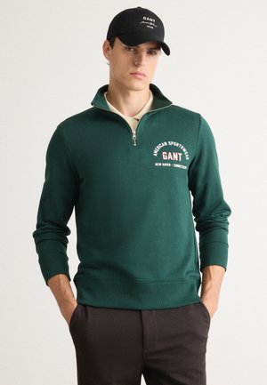 Muž v tmavozelenej mikine na zips s logom "GANT" a čiernou baseballovou čiapkou, stojí so zalomenými rukami v tmavých nohaviciach.