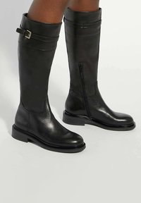 Black leather knee-high boots with a sleek design, featuring a side zipper, rounded toe, and a chunky rubber sole. Accented with a strap and buckle.