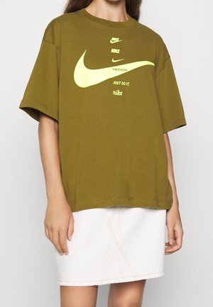 Nainen, jolla on päällään oliivinvihreä ylisuurikokoinen Nike t-paita, jossa on keltaiset logot ja valkoinen hame, yksiväristä taustaa vasten.