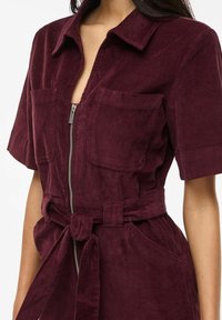 Tuta di velluto a coste color bordeaux con maniche corte, due tasche sul petto, chiusura frontale con cerniera e cintura annodata in vita.