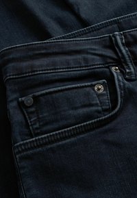 Donkerblauwe spijkerbroek met een klein voorvakje, metalen klinknagels en nette stiksels langs de naden. Zachte textuur met een lichte glans.