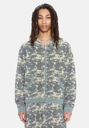 Personne portant un sweat à capuche zippé et un pantalon assortis en camouflage numérique, debout face à l'avant sur un fond uni.