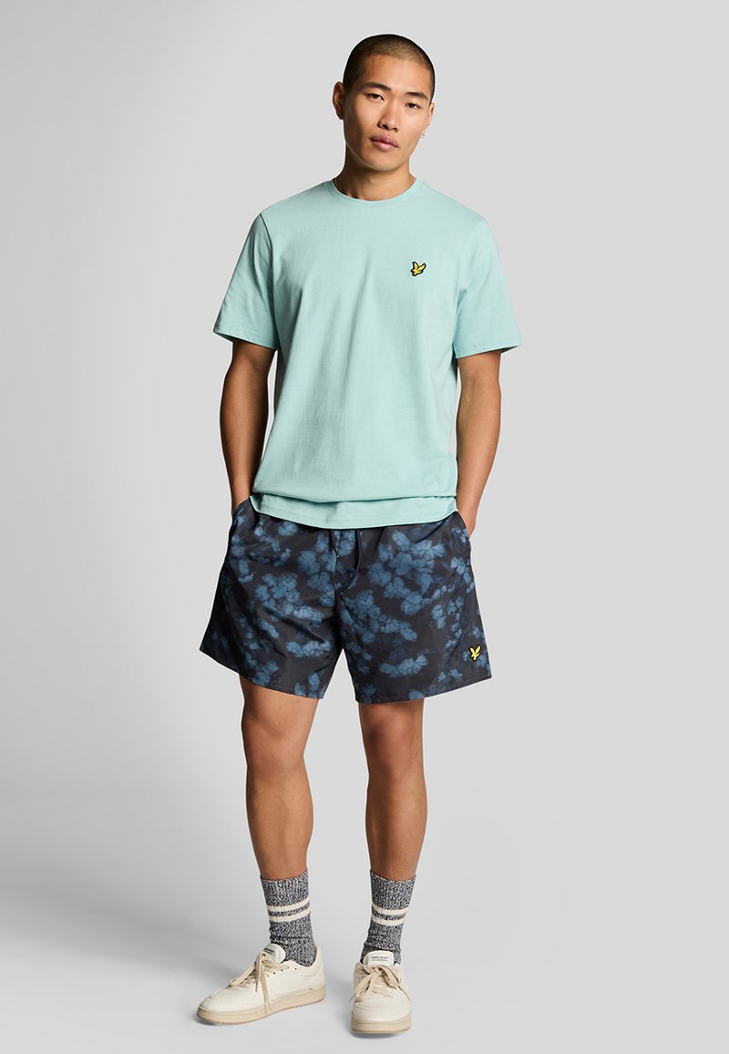 T-shirt di cotone azzurro chiaro con piccolo logo giallo, abbinato a pantaloncini da bagno blu scuro a motivo e sneakers crema con calzini a strisce grigie.