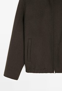 Veste en laine marron foncé avec un ourlet arrondi, fermeture éclair et poches latérales. Le tissu présente une finition texturée sans motifs.