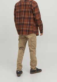 Hnědá kostkovaná flanelová košile s dlouhými rukávy, kombinovaná se světle béžovými cargo kalhotami s bočními kapsami a černými teniskami s hnědými podrážkami.