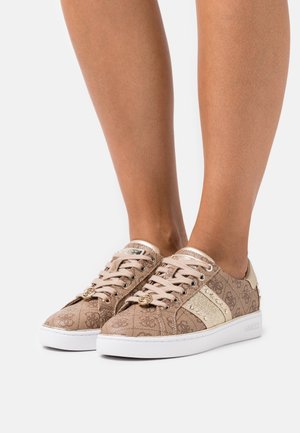 Beige Sneakers mit strukturiertem Obermaterial, ausgestattet mit goldenen Akzenten, einem glänzenden Streifen und einem Logo-Anhänger. Weiße Sohle mit dezenten Details.