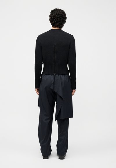 Άνδρας με σγουρά μαλλιά, γυρισμένος με την πλάτη, φοράει μαύρο πουλόβερ με φερμουάρ και σκούρο παντελόνι με ριγέ λεπτομέρειες και ασύμμετρο ύφασμα.