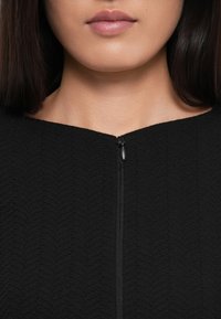 Onderkant van het gezicht en de nek van een vrouw die een zwart, getextureerd, rits-top draagt met een zichtbare ritstrekkertje in het midden van het sleutelbeen.