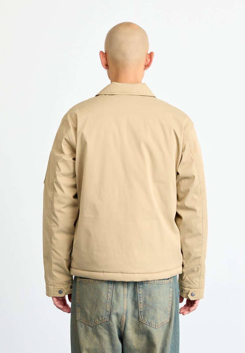 Homme avec la tête rasée portant une veste beige et un jean bleu se tient tourné vers l'arrière devant un fond blanc uni.
