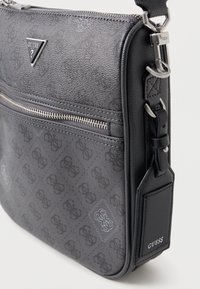 Šedá syntetická taška přes rameno s texturovaným povrchem, zipovou kapsou, logem GUESS a nastavitelným popruhem. Obsahuje jemný vzor na materiálu.